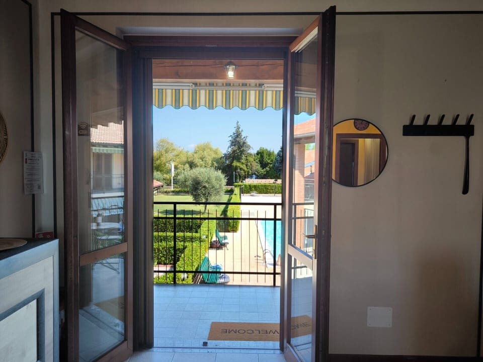 View from the living area of the apartment in a residence in Peschiera del Garda. The French doors lead directly onto the balcony overlooking the communal swimming pool and the well-kept internal garden, offering a relaxing and bright atmosphere ideal for a lakeside vacation.