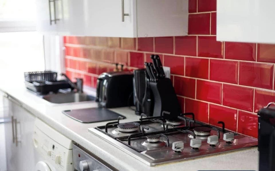 A modern kitchen with white cabinets, a washing machine, and a gas stove. The backsplash features glossy red tiles, adding a vibrant touch. Black utensils and a kettle sit neatly on the countertop for convenience and style.