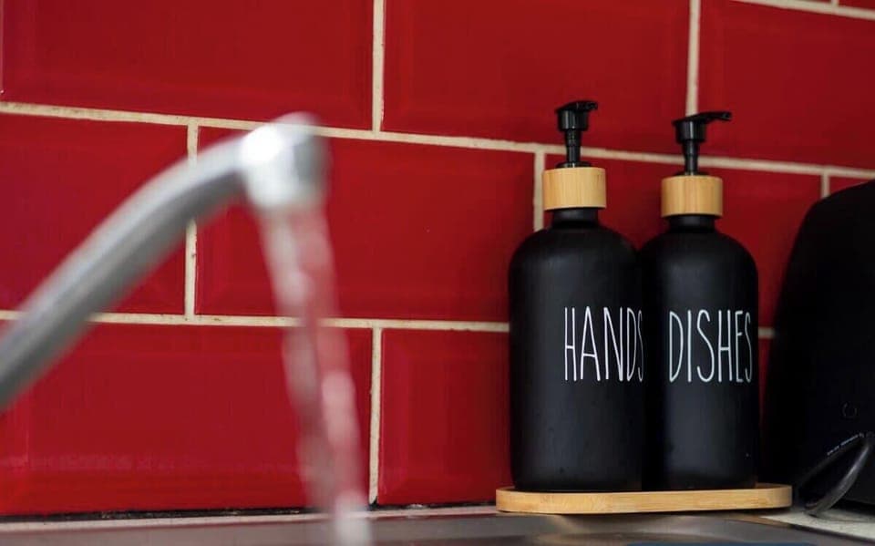 Water flows from a modern tap into a stainless steel sink. Two black pump bottles labelled "HAND" and "DISH" stand on the edge, set against a bold red tiled splashback, adding a stylish contrast to the kitchen scene.