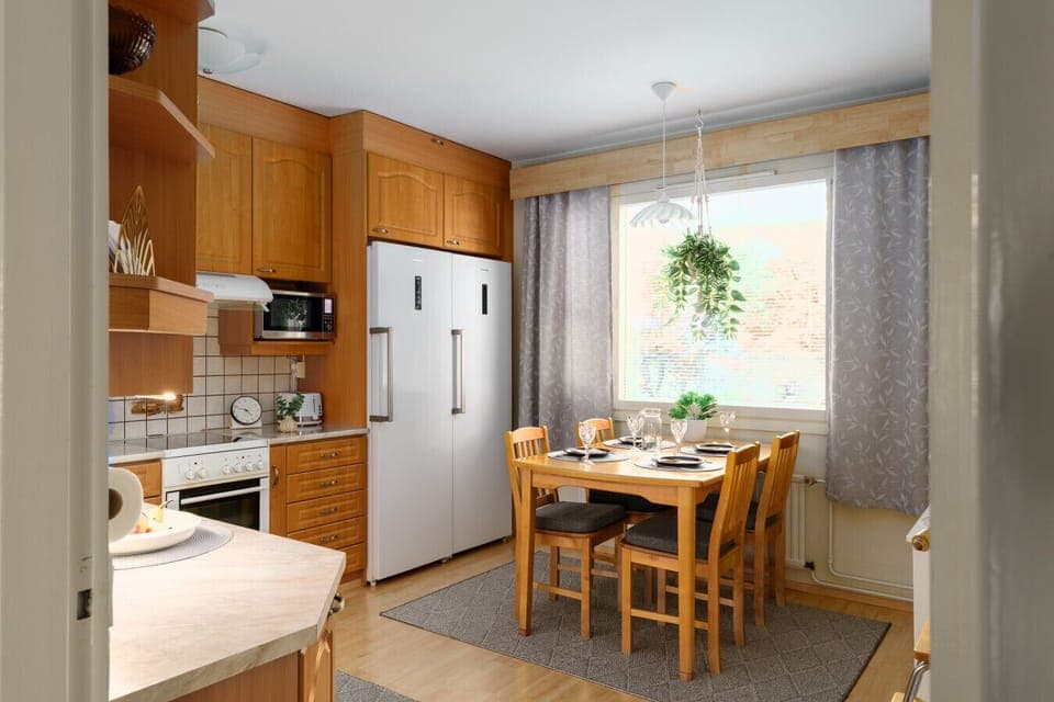 Bright and welcoming kitchen featuring warm wooden cabinetry and modern appliances including a double-door refrigerator, oven, microwave, and toaster. A cozy dining area with a wooden table and four chairs is set by the large window, allowing plenty of na