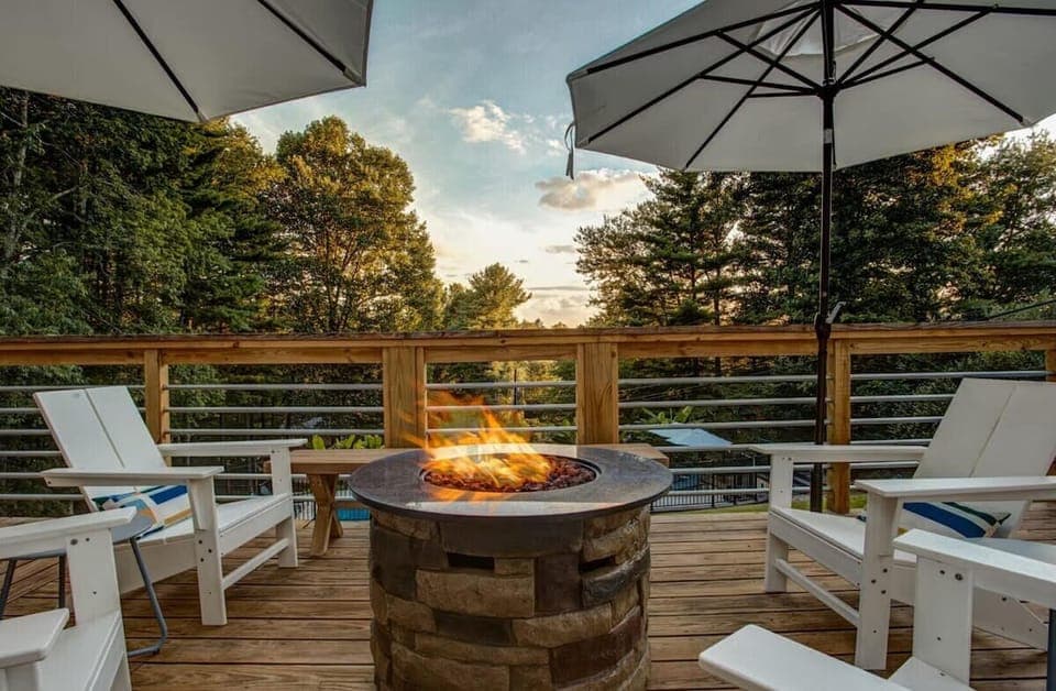 🔥 Cozy up around the stone fire pit with Adirondack seating 🪑 under large umbrellas ☀️, while enjoying serene forest views 🌲 and golden sunset skies 🌅.