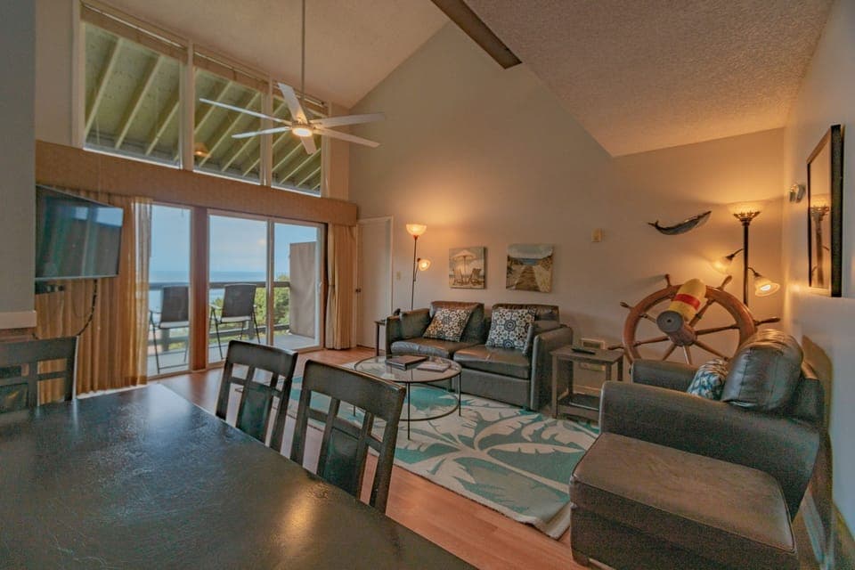 Spacious living area with vaulted ceiling, leather seating, and sliding doors opening to a balcony with ocean views.