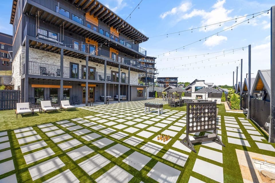 Spacious outdoor lounge area with modern turf and pavers, string lights, lounge chairs, oversized games, and shaded seating—perfect for entertaining or relaxing under the sun in a resort-style environment.