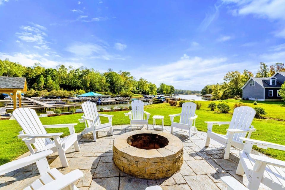 Fire Pit - White Adirondack chairs circle a stone fire pit on the patio, with the lake, dock, and pavilion just beyond.