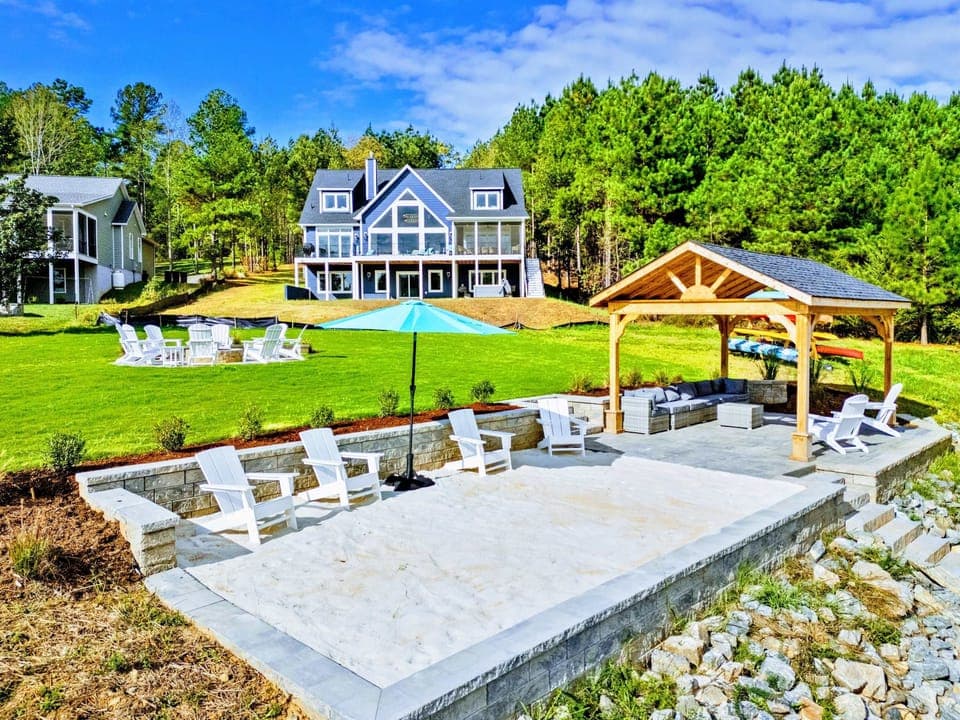 Lakeside Grounds - The full property from the waterfront, with Adirondack seating, a covered pavilion, and the home rising beyond a manicured lawn.