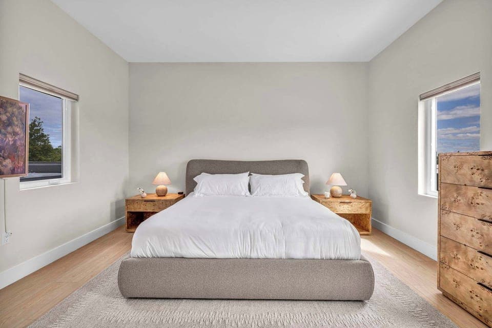 Wake up refreshed in a calming bedroom with a plush king bed, cozy lighting, and natural light from two windows for the perfect start to your day.