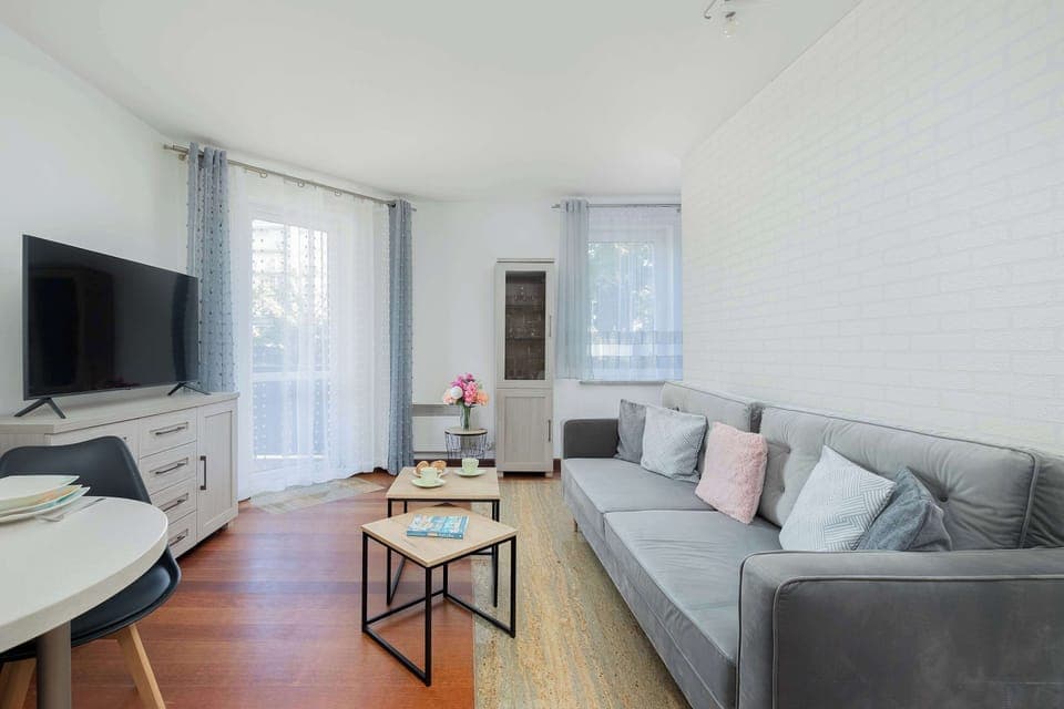A bright living room featuring a gray sofa, white walls, and large windows letting in plenty of light. A black coffee table and flat-screen TV complete the cozy modern setup.