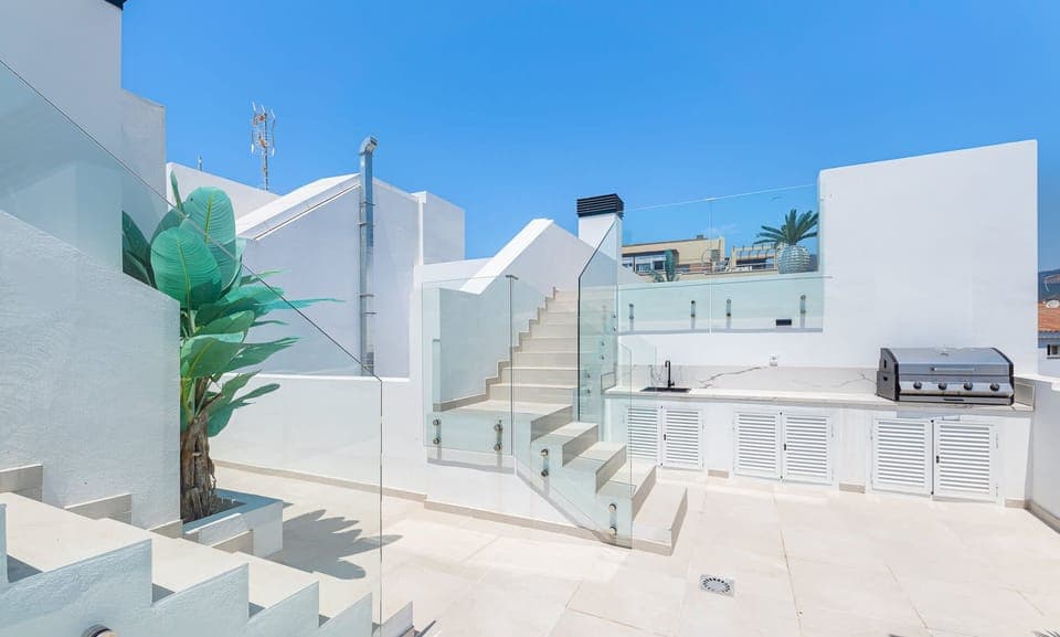 Rooftop outdoor kitchen area featuring built-in BBQ grill with white cabinet storage. Modern staircase with glass railings and large tropical plants. Clear blue sky with residential buildings in background.