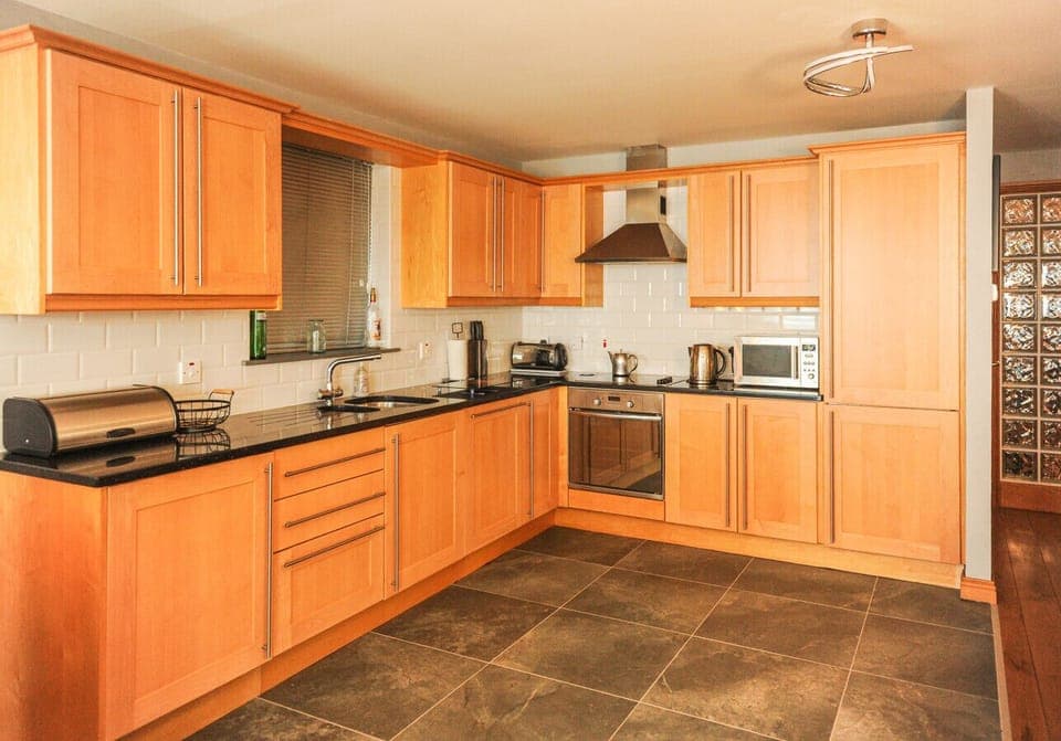 Fitted kitchen with light oak wall and base cabinets, dark worktops, white tile splashback, built-in oven, extractor hood, microwave, and kettle.