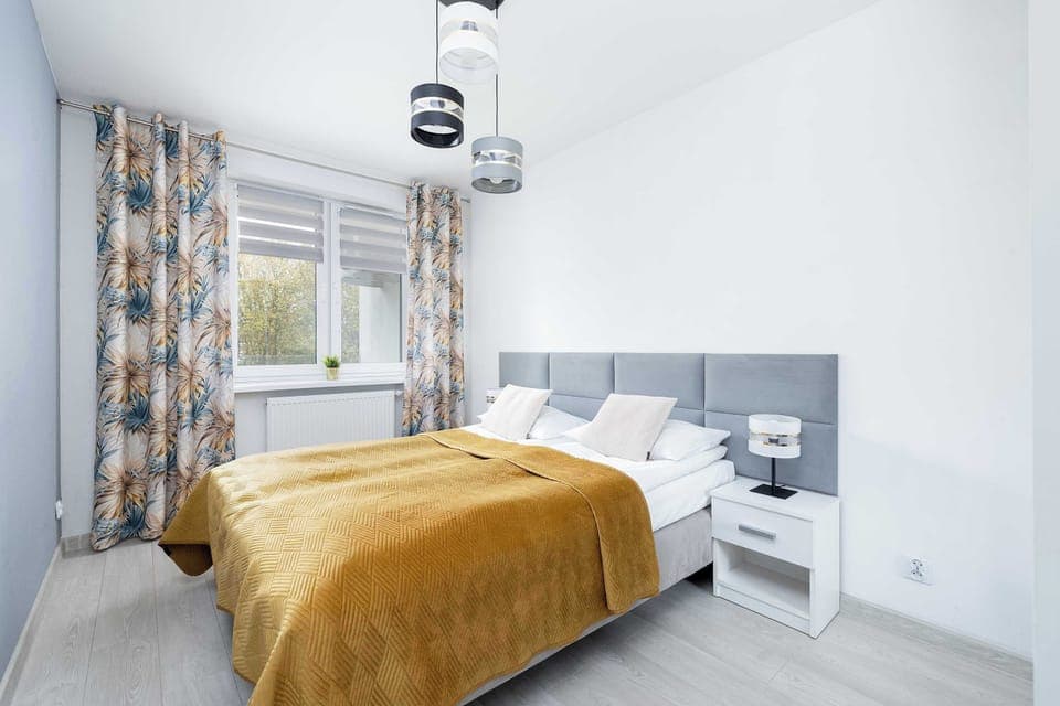 Bright bedroom with a comfortable double bed, golden blanket, and large windows bringing in natural light.