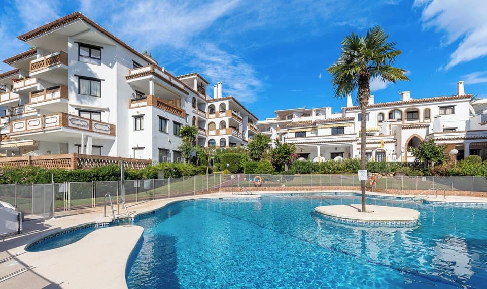 A sun-drenched view featuring a large swimming pool with inviting blue water, perfectly situated in front of a bright and airy apartment complex. Palm trees add a touch of tropical elegance to this idyllic setting.

