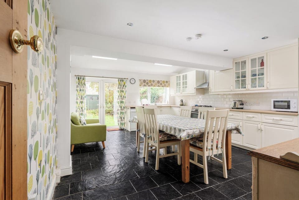 A bright, spacious kitchen with ample counter space, cabinet lighting, dishwasher, etc, and all essentials. Features a six-seater dining table, two-seater sofa, and double doors opening to the large garden—perfect for cooking, dining, and relaxing.