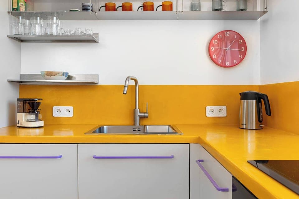 A compact kitchen with yellow countertops, white cabinets, and open shelving. The view through the window adds natural light to the space.