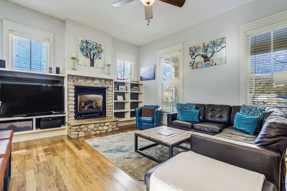 This bright living room features a stone fireplace, built-in shelving, and large windows that fill the space with natural light. Comfortable seating and a Smart TV create an inviting spot to unwind after a day exploring South Main.