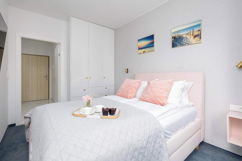 A bright bedroom with pastel decorations and a neatly made bed. Soft lighting and modern artwork enhance the serene atmosphere. The room looks clean and comfortable.