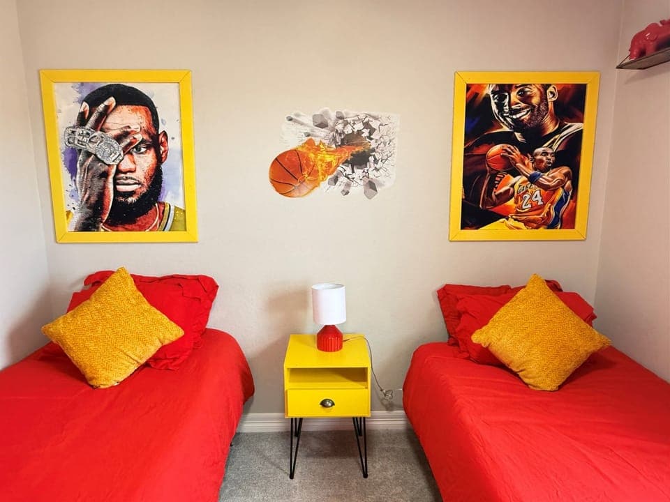 Closer look at the upstairs twin bedroom with two twin beds, a shared nightstand with a lamp between them, and sports themed artwork above.