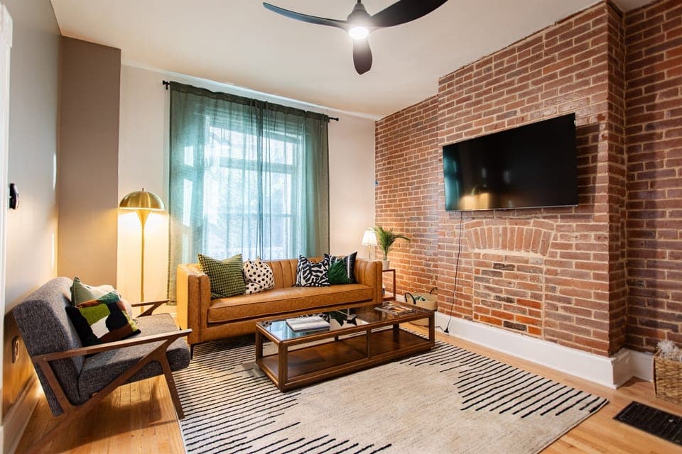 Cozy modern living room with exposed brick, mid-century furnishings, a mounted smart TV, and layered textures that create a warm, inviting space.