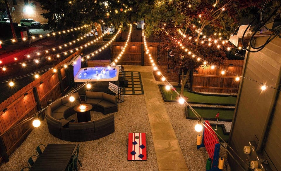 Backyard oasis with hot tub, fire pit lounge, mini golf, yard games, dining area, and enchanting string lights—perfect for relaxing, grilling, and unforgettable group fun