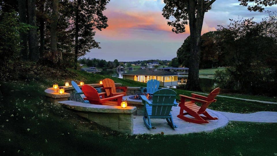 Lit fire-pit circle tucked into the hillside with Adirondacks in bright, joyful lagoons of color — sunset here feels like a ritual.