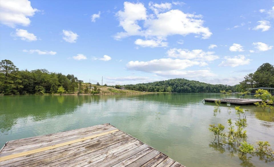 Peaceful lake with wooden dock surrounded by lush hills and clear blue skies in the surrounding area.