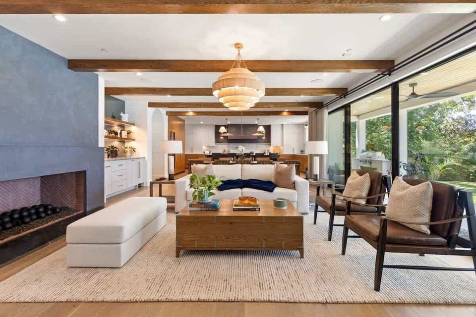This sophisticated living room showcases exposed wood beams, a striking fireplace, and expansive glass doors framing lake views. Plush seating, curated décor, and a statement chandelier create an elegant yet inviting atmosphere.