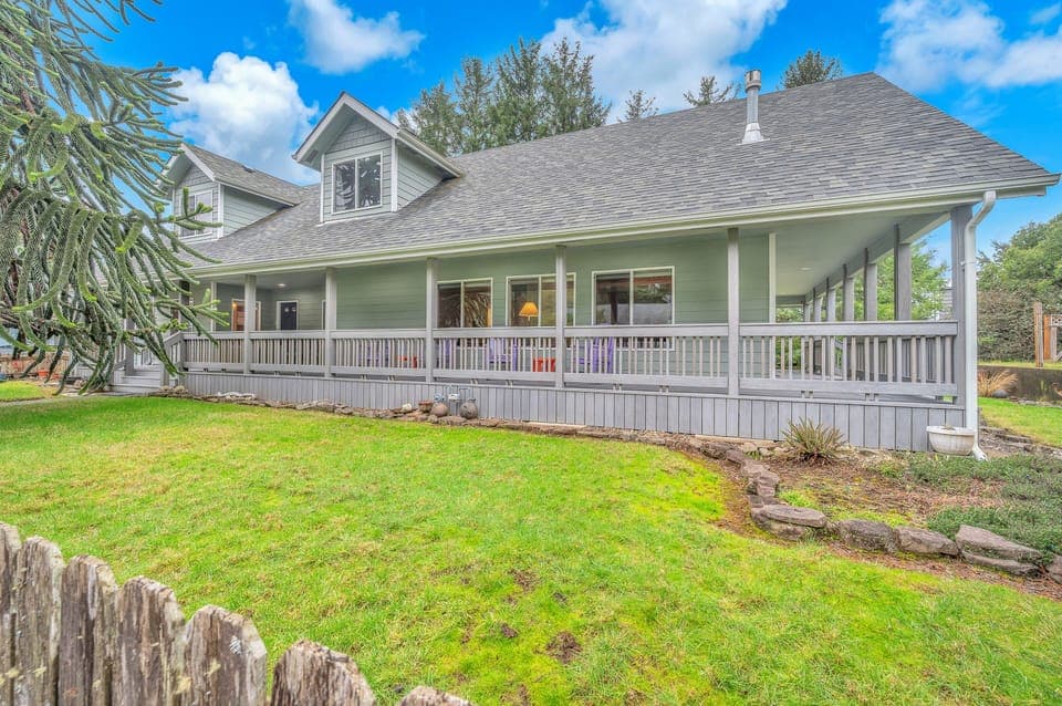 Charming green cottage with wraparound porch and spacious lawn offers peaceful retreat under blue skies.