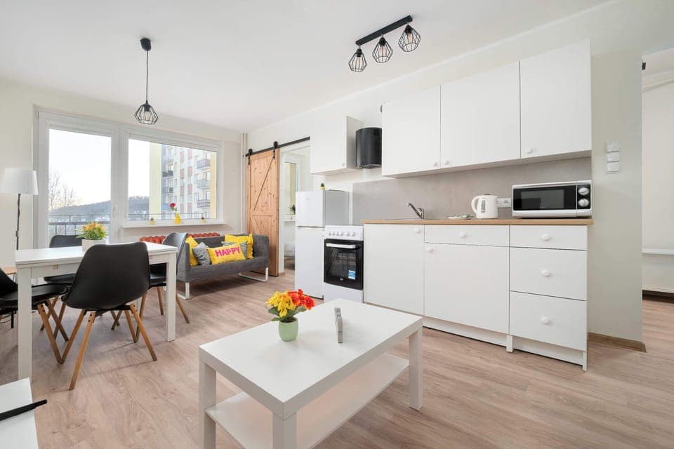 A wider angle of the open living area highlights a white coffee table and a compact dining space. Large windows bring in plenty of daylight, making the room feel airy and welcoming.
