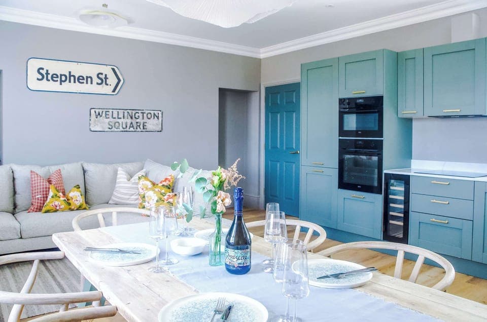 Contemporary living room with a plush beige sofa and patterned cushions, accented by quirky street-sign décor. Warm lighting and cloud-like fixtures add charm, creating a cozy, stylish space that flows into the kitchen and dining area.