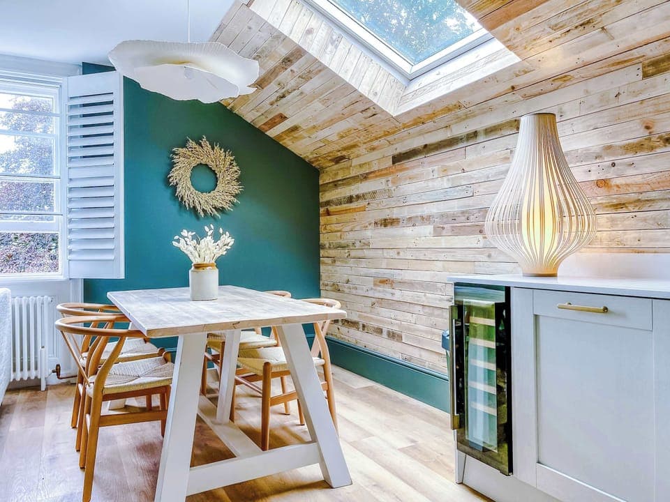 Dining area with Scandinavian charm, featuring a wooden table and chairs beneath a slanted ceiling with skylight. A green accent wall with sunburst mirror, sculptural lamp, and natural textures create a warm, modern, and inviting atmosphere.