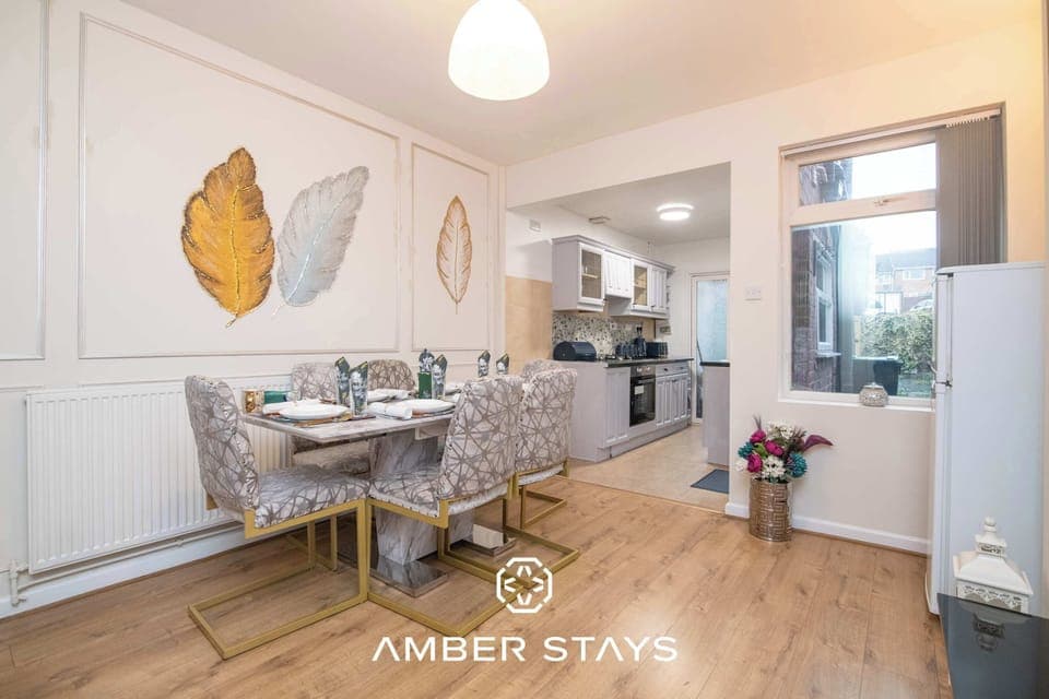 This bright dining area features gold-framed patterned chairs and elegant leaf artwork, flowing seamlessly into a light-filled kitchen. The neutral tones and wood flooring create a warm, inviting atmosphere.