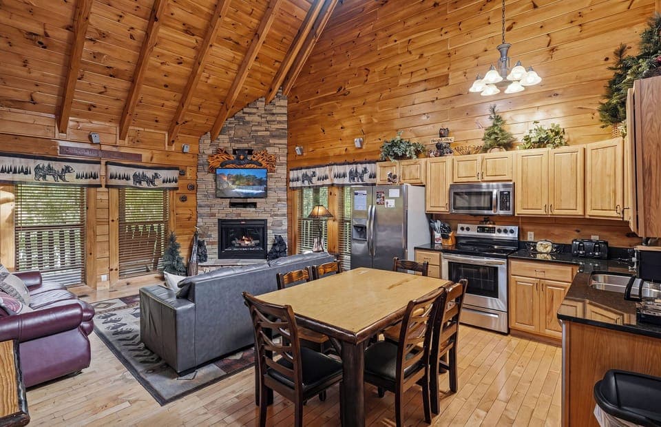Relax in your rustic cabin's open living space, where vaulted ceilings meet stone fireplace warmth and a fully-equipped kitchen awaits your culinary adventures.