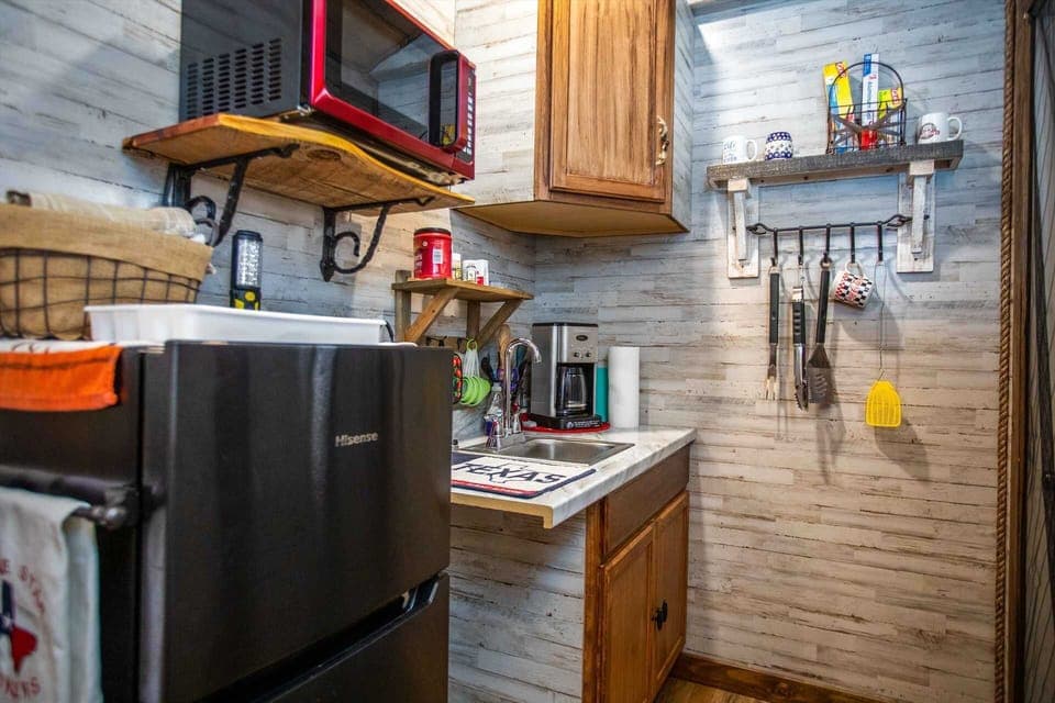 A clever and compact kitchenette area featuring a mini-fridge, a red microwave on a rustic wood shelf, and a sink. The space is accented with shiplap-style walls and handy storage for coffee and cooking essentials.