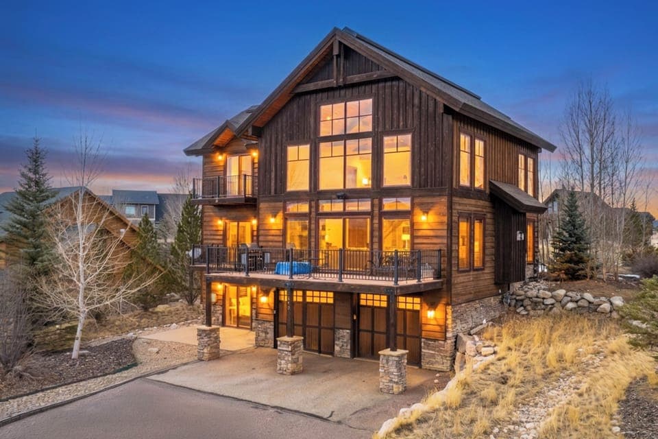 Stunning mountain lodge features warm wood exterior and expansive windows that glow invitingly at twilight in this peaceful neighborhood setting.