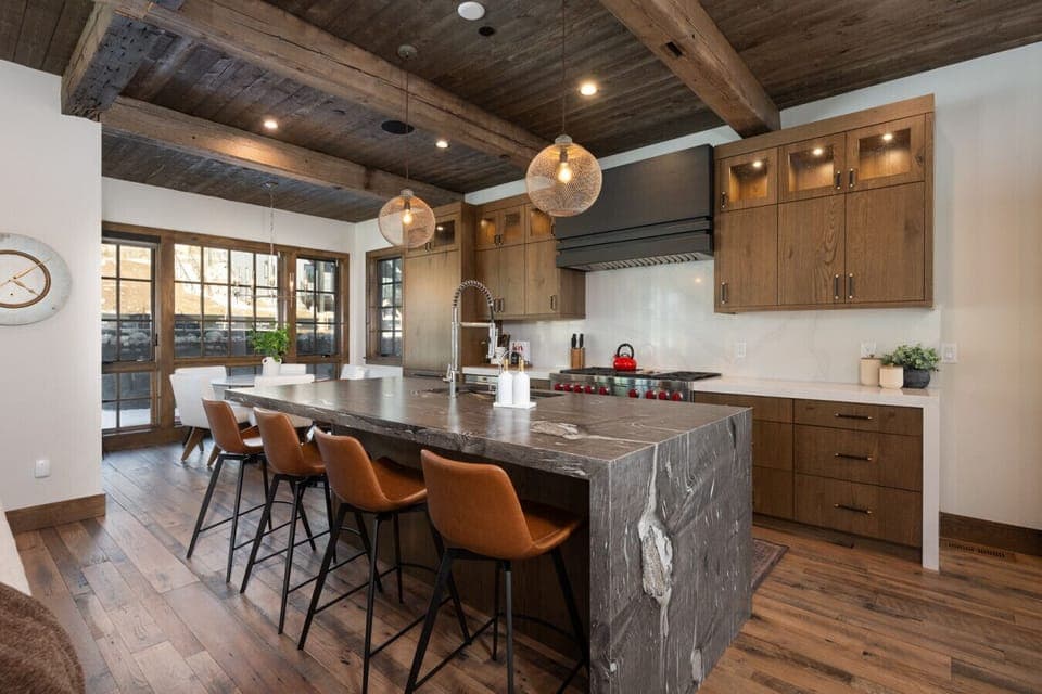 Open kitchen with large stone waterfall island, bar seating, wood cabinetry, and reclaimed beam ceiling