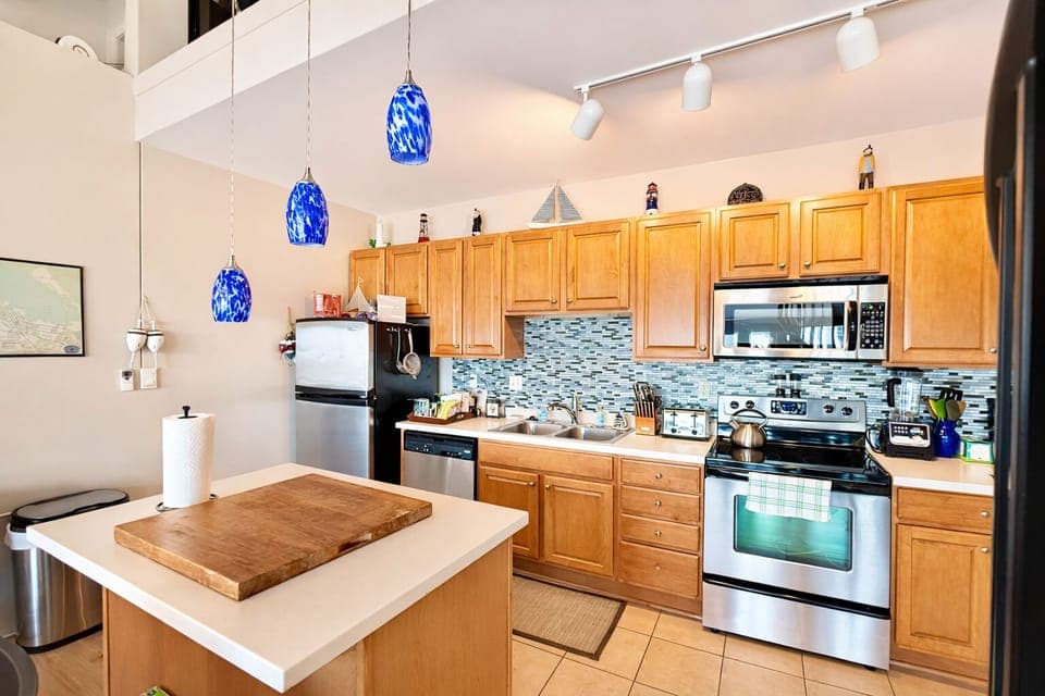 Bright, fully stocked kitchen with stainless steel appliances, coastal backsplash, and spacious island for meal prep or gathering. Blue pendant lighting adds charm to this open, loft-style cooking space.