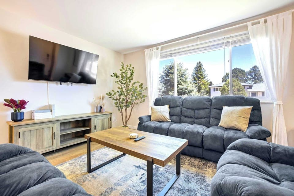 Welcome to Meadow Gate! The main level living room features a large wall-mounted TV, slate blue sofas, and a wood coffee table set on a patterned rug. Sheer curtains and wide windows keep the space bright and open throughout the day.
