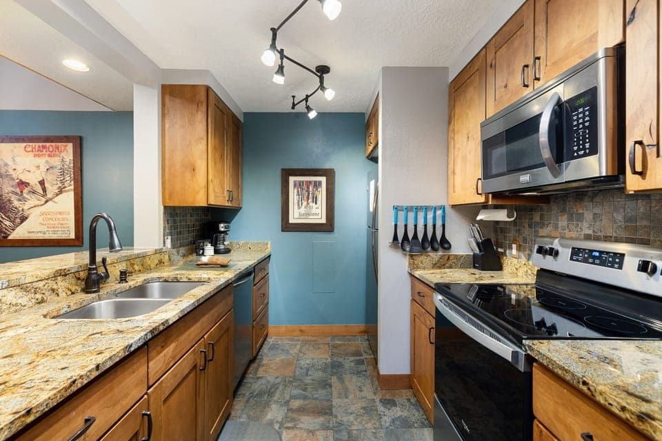 A modern kitchen with wood cabinets, granite countertops, a double sink, stainless steel appliances, and a tile floor. Track lighting is installed on the ceiling, and a framed picture hangs on the wall.