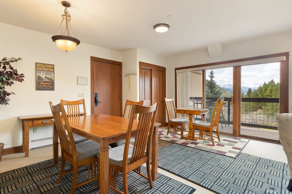 A dining area with a wooden table and six chairs. There is a wall-mounted light fixture, a small desk, and sliding glass doors leading to a balcony with a scenic outdoor view.