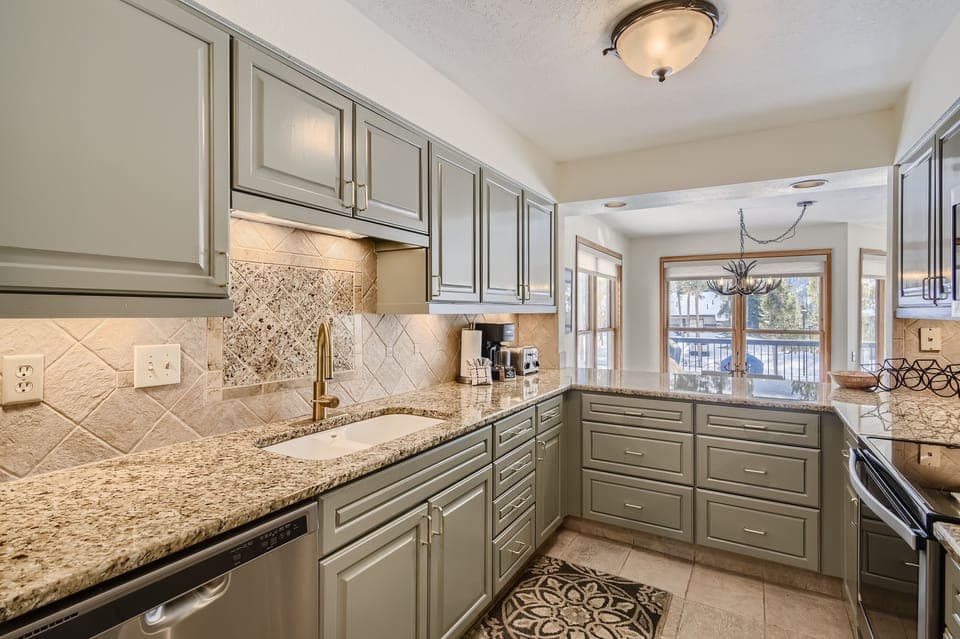 A modern kitchen with granite countertops, green cabinetry, stainless steel appliances, and a tile backsplash. A windowed dining area with a chandelier is visible in the background.