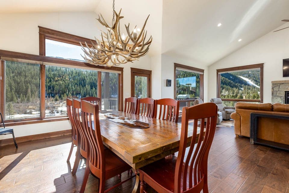 A dining room with a wooden table surrounded by wooden chairs. An antler chandelier hangs above the table. Large windows offer views of a forest outside. The room has hardwood flooring.