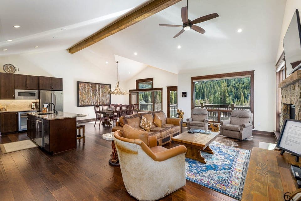 A spacious open-plan living area with wooden flooring, leather furniture, a kitchen with stainless steel appliances, and large windows offering a view of a forested landscape.