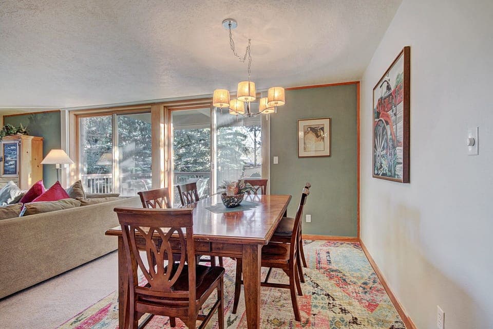 Dining room with a wooden table and chairs, a chandelier, and artwork on the wall. The adjacent living area has a sofa and large windows providing natural light with views of trees outside.