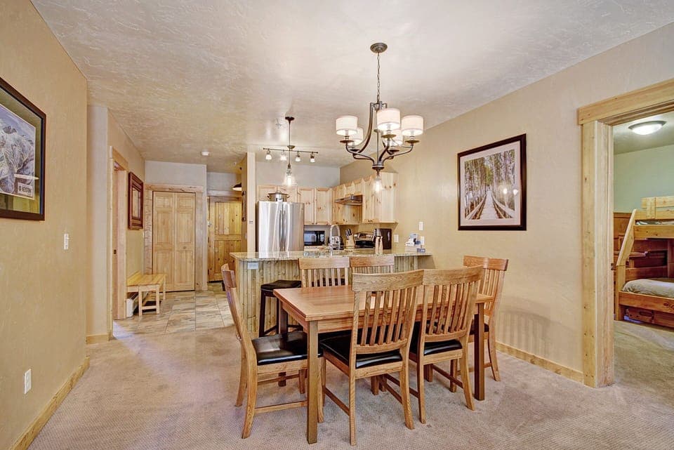 A cozy dining area with a wooden table and six chairs is adjacent to a bright kitchen. The room features beige walls, framed artwork, and modern light fixtures. A bunk bed is visible in the background.