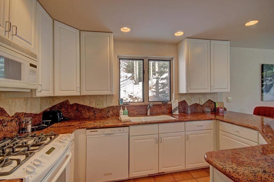 Modern kitchen with white cabinets, granite countertops, a dishwasher, stove, microwave, and sink. A window above the sink offers a view of trees and snow outside.
