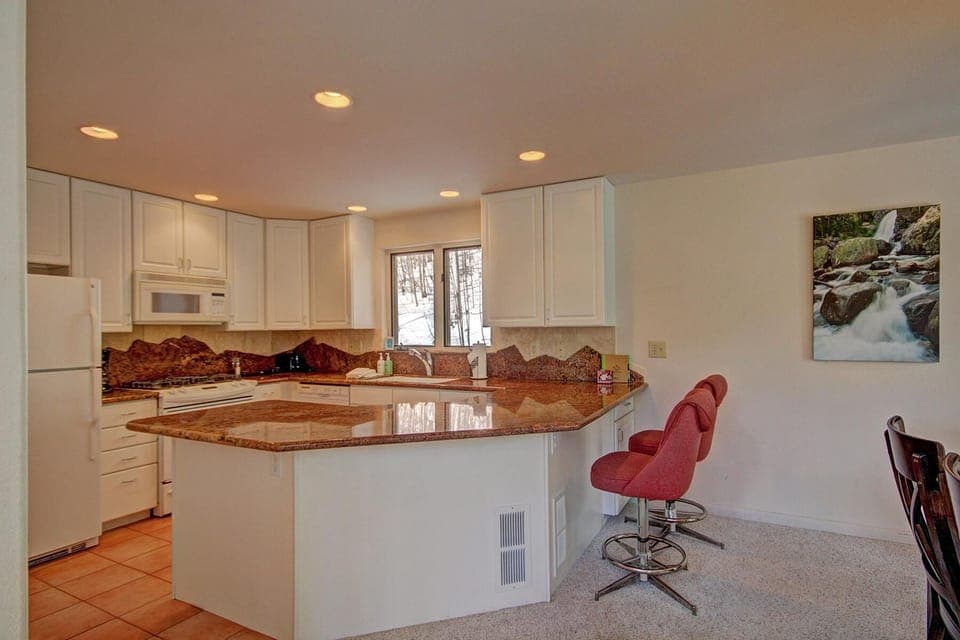A modern kitchen with white cabinets, granite countertops, a central island with bar stools, and recessed lighting. A window is above the sink and there is a picture of a waterfall on the wall.