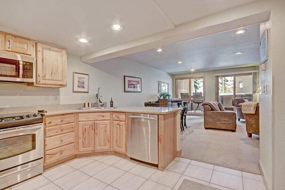 Open kitchen and living area with wooden cabinets, stainless steel appliances, and a dining table. Large windows provide natural light. Light-colored tile floors in the kitchen transition to carpet in the living area.