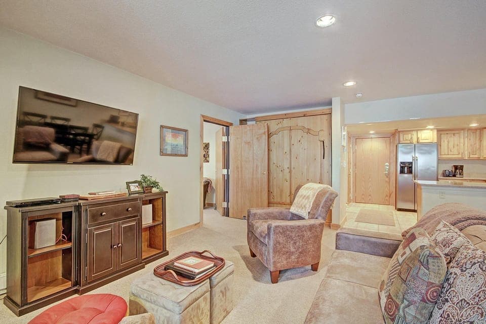 Cozy living room with a wall-mounted TV, brown armchair, plush couch, and a small cabinet. A doorway in the background leads to a kitchen with wooden cabinets and stainless steel appliances.