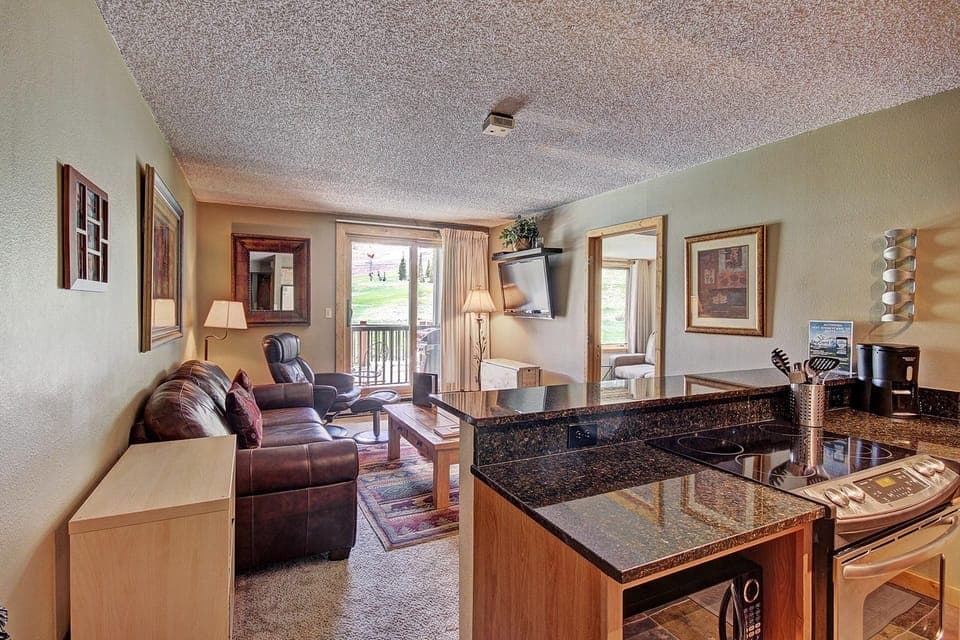 A cozy living area with a leather couch, framed artwork, and a wall-mounted TV. The room opens to a small balcony through sliding glass doors, offering a view of a grassy outdoor area.