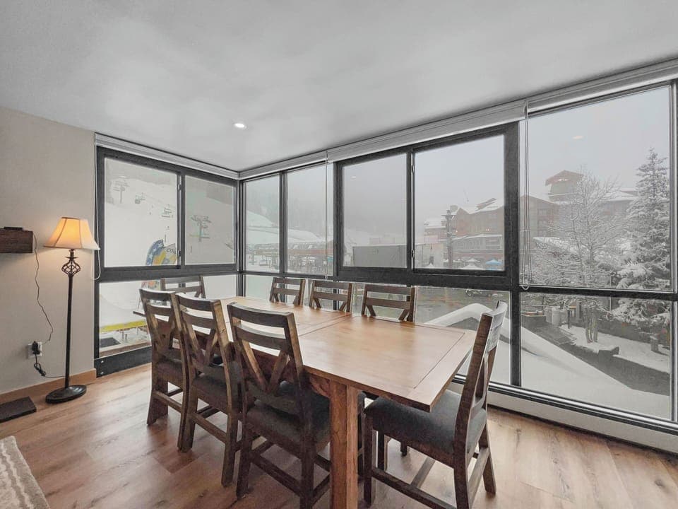 A dining room with a wooden table and eight chairs, large windows offering a snowy mountain view outside, a floor lamp in the corner, and wood flooring.