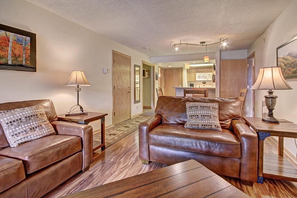 A cozy living room with brown leather sofas, wooden furniture, and a view into the kitchen with a bar counter and stools. Walls decorated with landscape art and warmly lit lamps.